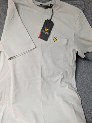 Ljusgrå t-shirt från Lyle & Scott - Ljusgrå t-shirt från Lyle & Scott med relaxed fit och liten bröstficka. Klassisk gul logga broderad på fickan. Tillverkad i mjuk bomull och har rund hals samt korta ärmar. Perfekt basic-plagg med stilren look.
