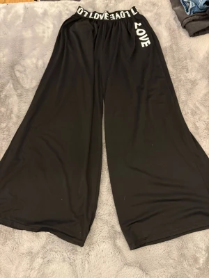 Svarta vida byxor från SHEIN - Säljer ett par svarta, supervida byxor från SHEIN med resår i midjan där det står 'LOVE' i vitt. Byxorna är gjorda i ett mjukt och stretchigt syntetmaterial och har en avslappnad passform. Perfekta för en chill och trendig look.