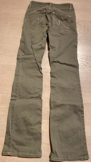 Olivgröna bootcut byxor från Nelly - Snygga olivgröna byxor från Nelly med bootcut passform. Byxorna har normal midja, klassisk femficksdesign och stängs med dubbla knappar och dragkedja. Perfekta till sneakers eller boots. Bra skick (knappt använda) men sliten färg fram pga miss i tvätten (mer vit se bild)