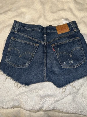 Levi's blå denimshorts med slitningar - Säljer ett par klassiska blå Levi's shorts i denim med råa, fransiga kanter och slitna detaljer framtill. Supersnygga, men säljer då storleken inte passar mig. 