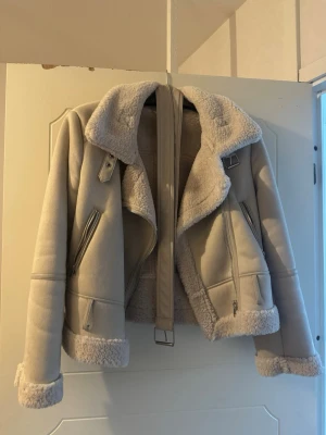 Beige ”fårskinnsjacka”/fodrad bikerjacka - Säljer en beige teddyjacka, mysigt foder och detaljer i form av dragkedjor och spänne vid kragen. Jackan har långa ärmar med uppvikta muddar och är perfekt för kyliga dagar. Snygg och trendig modell som ger en cool look. Man kan även stänga till jackan för en mer stilig look. 