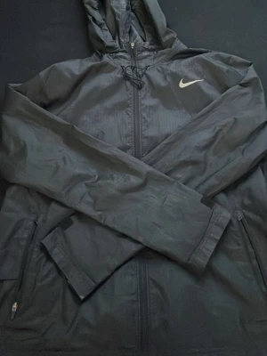 Svart vindjacka från Nike - Säljer en svart vindjacka från Nike med huva och dragkedja framtill. Jackan har en klassisk Nike-logga på bröstet, justerbar snörning vid huvan och fickor med dragkedja. Perfekt för blåsiga dagar och enkel att matcha med allt.