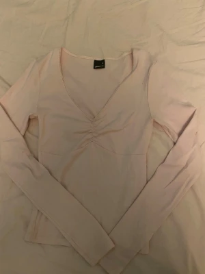 Ljusrosa v-ringad topp Gina Tricot - Supersöt ljusrosa topp från Gina Tricot i storlek XS. Modellen har v-ringning med rynkdetalj framtill och långa ärmar. Tunn och mjuk i materialet, perfekt till både jeans och kjol. Dekorativ kant runt halsen för extra detalj.