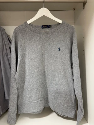 Grå tröja från Ralph Lauren - Säljer en stilren grå tröja från Polo Ralph Lauren med deras ikoniska logga broderad på bröstet. Tröjan har långa ärmar och en klassisk rund halsringning. Perfekt för en avslappnad och snygg look. Riktigt bra skick och inga defekter eller liknande👍Stickad grå tröja med blå logga den är storlek S men passar också större. Pris diskuterbart vid snabb affär! 