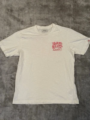 Vit graphic t-shirt från Replay med röd text - Snygg vit t-shirt från Replay med stor röd 'Replay Blue Jeans' print på ryggen och mindre logga på bröstet. Klassisk passform och rund halsringning. Tillverkad i mjuk bomull som känns skön mot huden. Perfekt för dig som gillar t-shirts med nåt litet extra på ocj tydliga loggor👌