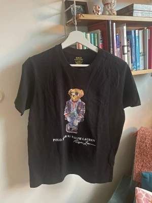 Svart Polo Bear t-shirt från Ralph Lauren - Hjälper nu brorsan att sälja en svart t-shirt från Polo Ralph Lauren med ikoniskt Polo Bear-tryck framtill. Modellen är custom slim fit och har rund hals. Tillverkad i mjuk bomull och passar perfekt för en cool och avslappnad stil.