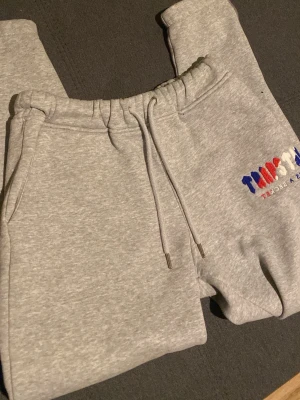 Grå mjukisbyxor från Trapstar - Säljer ett par grå mjukisbyxor från Trapstar med broderad logga i blått, rött och vitt på ena benet. Byxorna har dragsko i midjan och är gjorda i mjukt bomullsmaterial. Perfekta för chill eller streetwear.