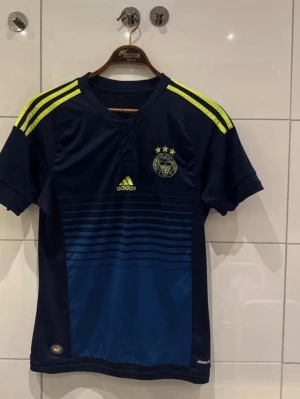 Fenerbahce Adidas matchtröja marinblå - Säljer en marinblå Fenerbahce matchtröja från Adidas med gula detaljer och tre stjärnor på bröstet. Tröjan har gula ränder på axlarna, diskret randigt mönster framtill och är tillverkad i Climacool-material som andas. Perfekt för dig som älskar fotboll och vill ha en snygg supportertröja.