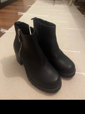 Svarta boots med klack och dragkedja - Säljer ett par helt nya svarta boots i skinnimitation med chunky klack och grov sula. Skorna har både guldfärgad och svart dragkedja på sidorna för en snygg kontrast. Rund tå och enkel, stilren design som passar till många outfits. 