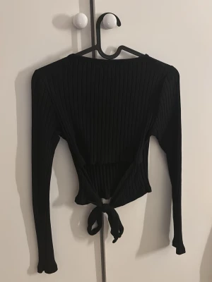 Svart ribbad topp med öppen rygg - Säljer en svart ribbad topp med lite öppen rygg från New Yorker i storlek XS. Toppen har långa ärmar, croppad passform och snygg knytning i ryggen. Den är stretchig. Gåva vid köp!🎁