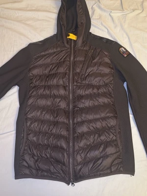 Svart pufferjacka från Parajumpers - Säljer en svart pufferjacka från Parajumpers med huva och gul detalj vid halsen. Jackan har quiltad framsida och baksida, stickade ärmar och sidopaneler samt dragkedja framtill och på fickorna. Märkets patch sitter på ena ärmen.