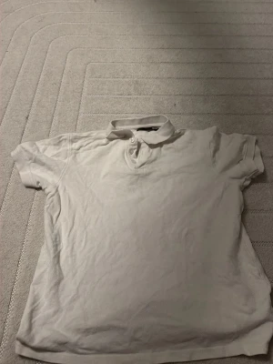 Vit kortärmad polotopp - Säljer en enkel vit polotopp med korta ärmar och klassisk krage. Toppen har knappar framtill och är gjord i mjukt bomullsmaterial. Perfekt till jeans eller kjol för en clean och stilren look.