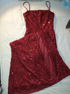 Vinröd glittrig långklänning - Säljer en vinröd långklänning med tunna axelband och massor av glittrande paljetter över hela klänningen. Klänningen har en hög slits framtill som ger en extra snygg touch. Perfekt för dig som vill sticka ut på bal eller fest! Öppen rygg. Använd endast 1 gång. Nypris 499kr. 