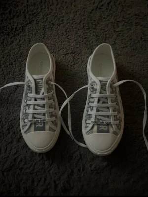 Christian Dior sneakers med monogram - Säljer ett par snygga Christian Dior sneakers i vitt och grått med klassiskt Dior-monogram. Skorna har snörning, rund tå och vit platt sula med stjärndetalj undertill. Tillverkade i textil och skinn, med logotyp på plösen och sidan. Perfekta för dig som vill ha något extra till din outfit.
