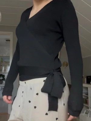 Svart omlottopp med knyt i midjan - Säljer en svart omlottopp med lång ärm och v-ringning. Toppen har ett brett knytband i midjan som ger en snygg siluett och är tillverkad i ett mjukt, stretchigt material. 