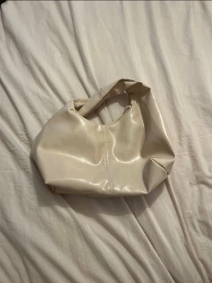 Beige axelväska  - Säljer en stilren beige axelväska med mjuk form och bred axelrem. Perfekt för dig som vill ha en enkel men trendig väska till vardagen. Väskan har en snygg söm i mitten och rymmer det viktigaste.