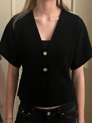 Svart blus med guldknappar  - Säljer en svart blus från missäy med v-ringning och två dekorativa guldknappar framtill. Modellen är croppad med korta, vida ärmar och rak passform. Perfekt att styla med både jeans och kjol för en clean look.