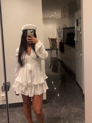 Studentklänning/Vit klänning - Säljer min vita studentklänning som jag köpte till studenten men tyvärr aldrig använde. Den är alltså helt oanvänd, i perfekt skick och redo för någon annans stora dag 🤍  Klänningen sitter otroligt fint på kroppen med markerad midja, v-ringning och ett knytband som gör att man kan justera passformen precis som man vill. Kjolen har flera lager med volanger som ger ett jättefint, luftigt fall och känns både elegant och somrig. Tyget är lätt och bekvämt – perfekt för en lång studentdag. 