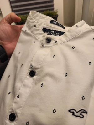 Vit Hollister henley med svarta knappar S - Säljer en vit henley från Hollister med svarta knappar och diskret svartmönstrade små rutor över hela skjortan. Skjortan har muscle fit-passform, rund krage och Hollister-logga broderad på bröstet. Tillverkad i stretchig bomull för extra komfort.