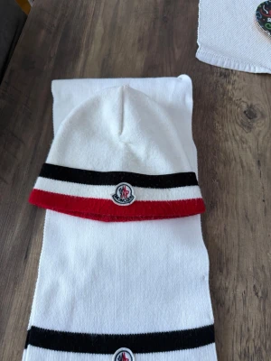 Vit Moncler mössa och halsduk - Säljer ett snyggt set från Moncler med vit mössa och matchande halsduk. Båda har svarta och röda ränder samt Moncler-logga framtill. Perfekt accessoar för kyliga dagar och ger en clean look.