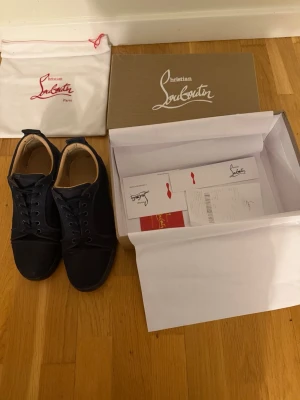 Mörkblå sneakers från Christian Louboutin - Snygga mörkblå sneakers från Christian Louboutin i mocka med klassisk röd sula och diskret broderad logga på hälen. Skorna har rund tå, snörning och beige innersula. Kommer med originalkartong och dustbag. 