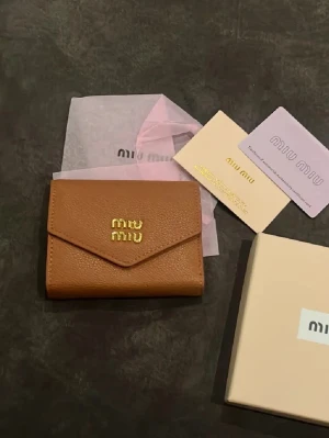 Brun plånbok från Miu Miu i skinn - Säljer en snygg brun plånbok från Miu Miu i äkta skinn. Plånboken har guldfärgad logga framtill, flera kortfack och ett separat myntfack. Kommer med original ask och certifikat. Perfekt storlek för att ha med sig överallt.