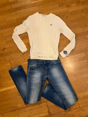 Vit kabelstickad tröja Morris - Säljer en vit kabelstickad tröja från Morris i storlek M. Tröjan har rund hals, ribbade muddar och en snygg broderad logga på bröstet. Perfekt för dig som gillar klassisk och stilren look.   Modellen är 182