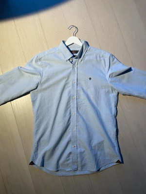 Ljusblå skjorta från Morris - Nypris är 1300kr. Säljer en stilren ljusblå skjorta från Morris med klassisk krage och knappar hela vägen. Skjortan har lång ärm och en liten broderad logga på bröstet. Perfekt för dig som gillar enkel och clean stil.
