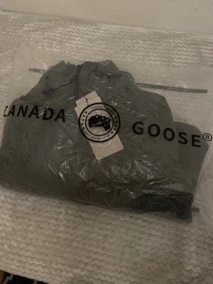 Mörkgrön pufferjacka från Canada Goose - Säljer en mörkgrön pufferjacka från Canada Goose med huva och svarta detaljer. Jackan har dragkedja framtill, två sidofickor och en patch med logga på ärmen. Perfekt för kalla dagar och riktigt snygg till streetwear-stilen.