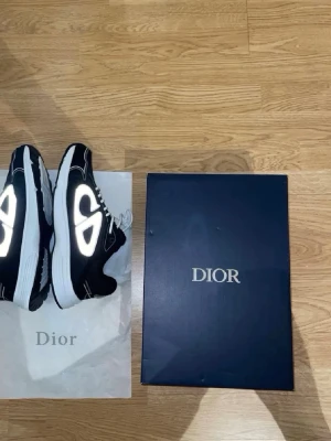 Svarta Dior B30 sneakers - Säljer ett par svarta Dior B30 sneakers med vita reflekterande detaljer och vita snören. Skorna har en chunky vit sula och meshpaneler för extra komfort. Märket syns tydligt på plösen och innersulan. Riktigt snygga och trendiga sneakers för dig som gillar lyxiga detaljer.