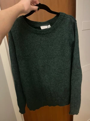 Mörkgrön stickad tröja från Vila - Mysig mörkgrön stickad tröja från Vila med rund halsringning och långa ärmar. Tröjan har ribbade muddar vid ärmslut, hals och nederkant. Perfekt att slänga på sig när det är kyligt ute.