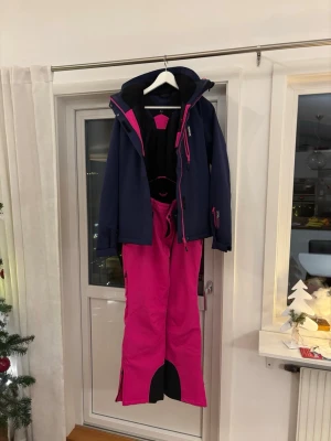 Rosa/lila skidset från Nanok  - Säljer ett skidset med skidjacka och skidbyxor  från Nanok i en riktigt snygg rosa/lila färg med svarta detaljer och byxorna har praktiska hängslen. Byxorna har dragkedjor vid bensluten och vid midjan och är perfekta för vinterns alla äventyr. Jackan har skidlås, ficka för liftkort och luva. Passar dig som vill sticka ut i backen! Skicka för fler bilder!