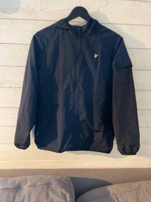 Svart vindjacka från Lyle & Scott - Säljer en svart vindjacka från Lyle & Scott med huva och dragkedja framtill. Jackan har det klassiska Lyle & Scott märket framtill och en ficka på ena ärmen. Perfekt för blåsiga dagar och enkel att matcha. Dm för frågor. 