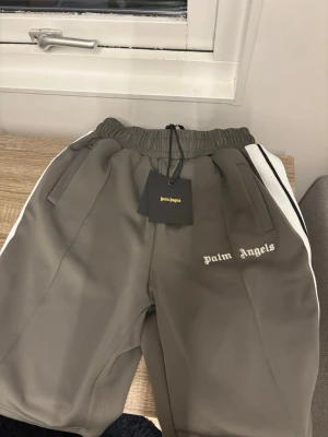 Palm Angels grå shorts - Snygga grå shorts från Palm Angels med vita ränder längs sidorna och logga broderad framtill. De har elastisk midja med snörning och är tillverkade i ett mjukt, sportigt polyestermaterial. Perfekta för en chill och trendig look.