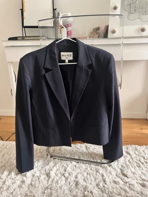 Mörkblå kavaj från NA-KD, strl 34 - Snygg mörkblå kavaj från NA-KD i croppad modell med krage och knapp framtill. Rak passform och långa ärmar, perfekt till både jeans och kjol. Tillverkad i ett mjukt material som känns bekvämt hela dagen. Lite smutsig runt halsen, annars som ny.