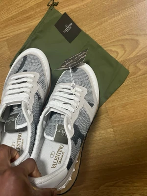 Valentino Garavani Rockrunner sneakers - Snygga Valentino Garavani Rockrunner sneakers i vitt, grått och svart med camouflagemönster. Skorna har mesh och mockadetaljer, vita snören och coola nitar på sulan. Perfekta för dig som vill ha en unik och trendig look.