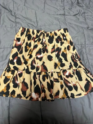 Leopardmönstrad kortkjol från SHEIN - Säljer en kort kjol från SHEIN med leopardmönster i beige, svart och bruna toner. Kjolen har volangkant och resår i midjan med knytband , storlek XS