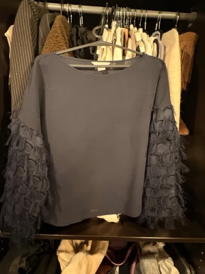 Mörkblå blus med fransiga ärmar H&M - Unik mörkblå blus från H&M i storlek 32. Toppen har vida, långärmade ärmar med fransiga detaljer och prickar i tyget som ger en cool struktur. Rak passform och rund halsringning. Perfekt för dig som vill sticka ut lite extra.
