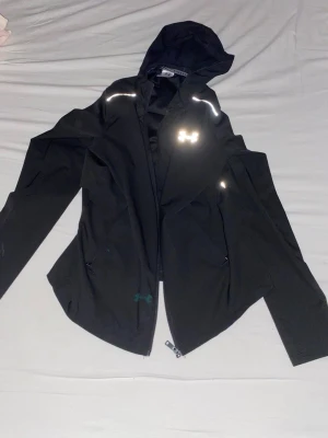 Svart vindjacka från Under Armour - Svart vindjacka med huva från Under Armour. Jackan har reflekterande detaljer, dragkedja framtill och logga på bröstet samt stor vit logga på ryggen. Tillverkad i lätt polyester, perfekt för aktiva dagar och träning utomhus.