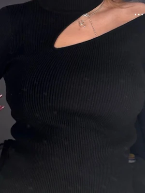 Svart ribbad polotopp med cut out - Säljer en svart ribbad polotopp med en snygg cut out-detalj vid axeln. Toppen är tight och har lång ärm, vilket ger en clean och trendig look. Perfekt att styla med smycken för en extra touch.