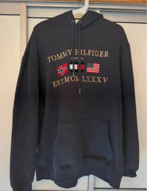 Svart hoodie Tommy Hilfiger - Svart hoodie från Tommy Hilfiger med tryck av flaggor och guldfärgad text på bröstet. Tröjan har huva med snörning och en stor magficka. Perfekt för dig som gillar klassisk streetstyle och vill ha en bekväm och snygg look.
