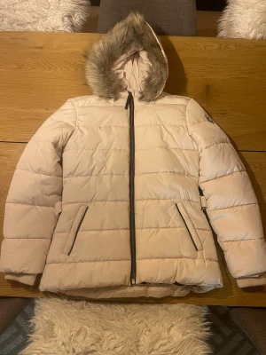 Beige vinterjacka från Garcia - Snygg beige vinterjacka från Garcia med huva och fuskpäls. Jackan har vattenavvisande material, svarta dragkedjor och två fickor framtill. Perfekt för kalla dagar med sin quiltade design och ribbade muddar vid ärmslut. Stilsäker look för vintern! Helt oanvänd, nypris 800kr