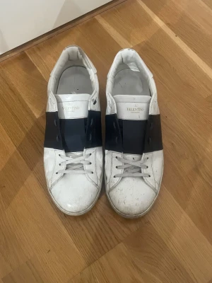 Valentino Garavani Open sneakers - Säljer ett par Valentino Garavani Open sneakers i vitt skinn med bred svart läderdetalj över mitten. Klassisk rund tå, platt sula och snörning framtill. Snygg och stilren design som passar till jeans eller dressade outfits. Storlek 46. Lite smutsiga men annars i fint skick. Endast skorna medföljer.
