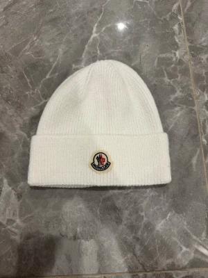 Vit stickad Moncler mössa - Snygg vit mössa från Moncler med klassisk ribbstickad design och uppvikt kant. Mössan har Moncler-loggan framtill och är tillverkad i mjukt stickat material som håller dig varm och stilren under kalla dagar.