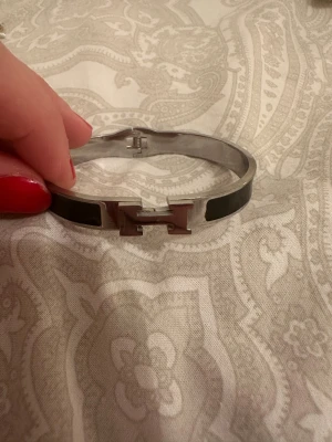 Ingen väst på bilderna - Bilderna visar ett armband med H-formad detalj i metall och svarta inslag, ingen väst syns på bilderna.