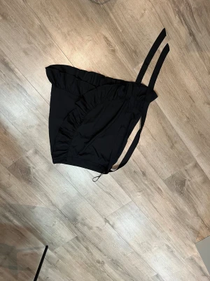 Svart omlott kjol med volang från Angeleye & Me - Säljer en svart kjol från Angeleye & Me i storlek S. Materialet känns lätt. Knytband i sidan för att justera passformen. Helt oanvänd.