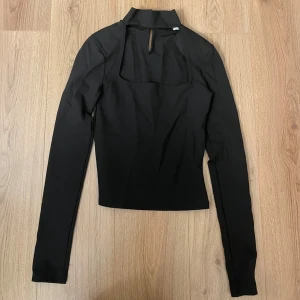 Svart långärmad topp - Snygg svart topp med lång ärm och cool cut-out detalj framtill. Toppen har en hög krage och knappstängning i nacken. Materialet känns mjukt och stretchigt, perfekt för att skapa en clean och stilren look. Oanvänd 