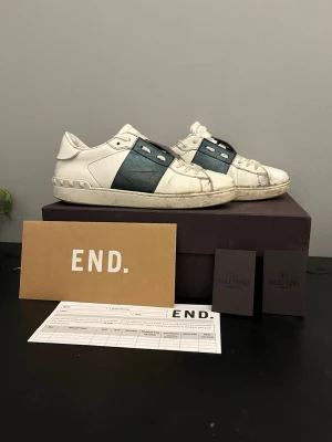 Valentino Garavani sneakers vit/blå - Säljer ett par Valentino Garavani sneakers i vitt skinn med bred mörkblå rem över sidan. Skorna har snörning, platt sula och rund tå. Märkeslogga under sulan och detaljerade sömmar. Perfekt för dig som gillar exklusiva och stilrena sneakers.