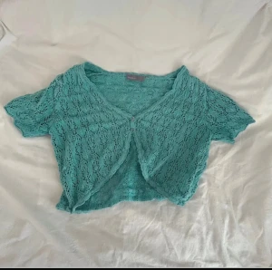 Turkos bolerotopp med spetsdetaljer - Säljer en turkos bolerotopp från Vero Moda med korta ärmar och v-ringad hals. Toppen har spetsmönster och två små knappar framtill. Croppad passform som ger en cool och trendig look. Perfekt för att lyfta din outfit med lite färg.