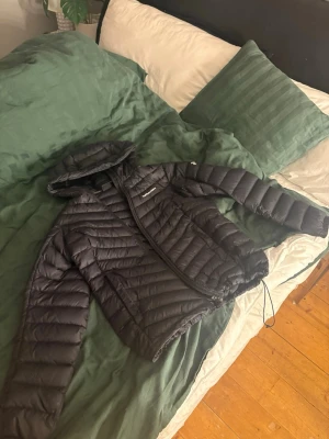 Peak Performance Jacka - Svart dunjacka från Peak Performance i storlek L. Jackan har huva, dragkedja och snyggt quiltad design. Perfekt för kyliga dagar och har en stilren look med logga på bröstet. Passar dig som vill ha en varm och lätt jacka med sportig vibe. Inga skador, kanske nån fläck nånstans men inget ja sett. Pris kan diskuteras 